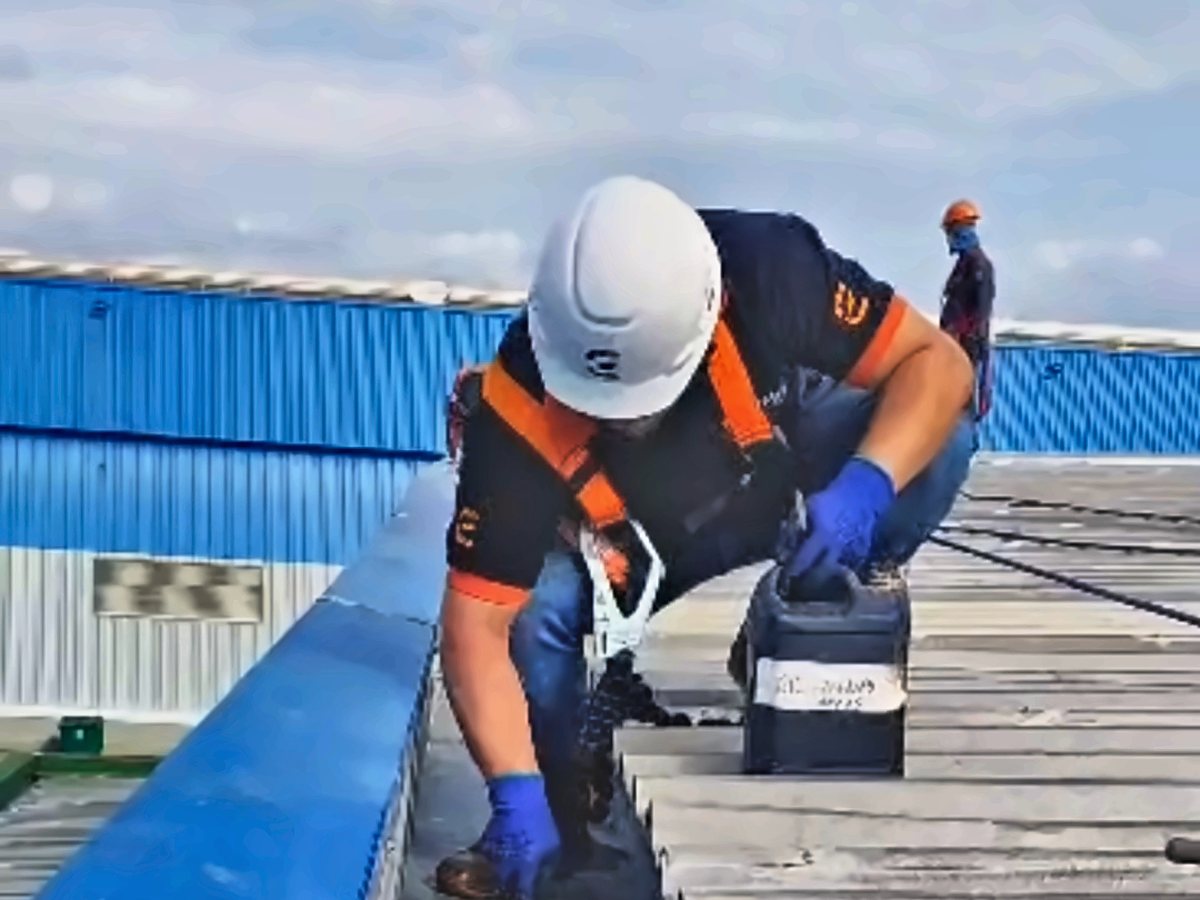 Profissional com cinto de segurança, capacete e luvas fazendo a manutenção de uma calha na cobertura de um galpão